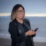 Picture is a headshot of Monica, creator of Portrait of a fangirl, with the beach behind her. She is holding her cellphone in her hands as if she is texting