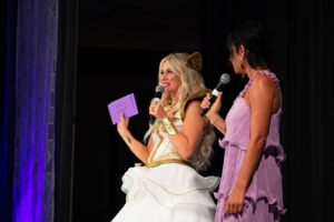 Ashley Eckstein stands on stage at the Her Universe Fashion Show, wearing a She-Ra-inspired gown.