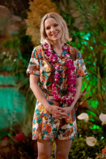 Eleanor stands with her hands clasped and smiling at someone who is not seen in this photo. She is wearing a Hawaiian themed romper and a floral lei around her neck. There are tropical flowers in the distance. 