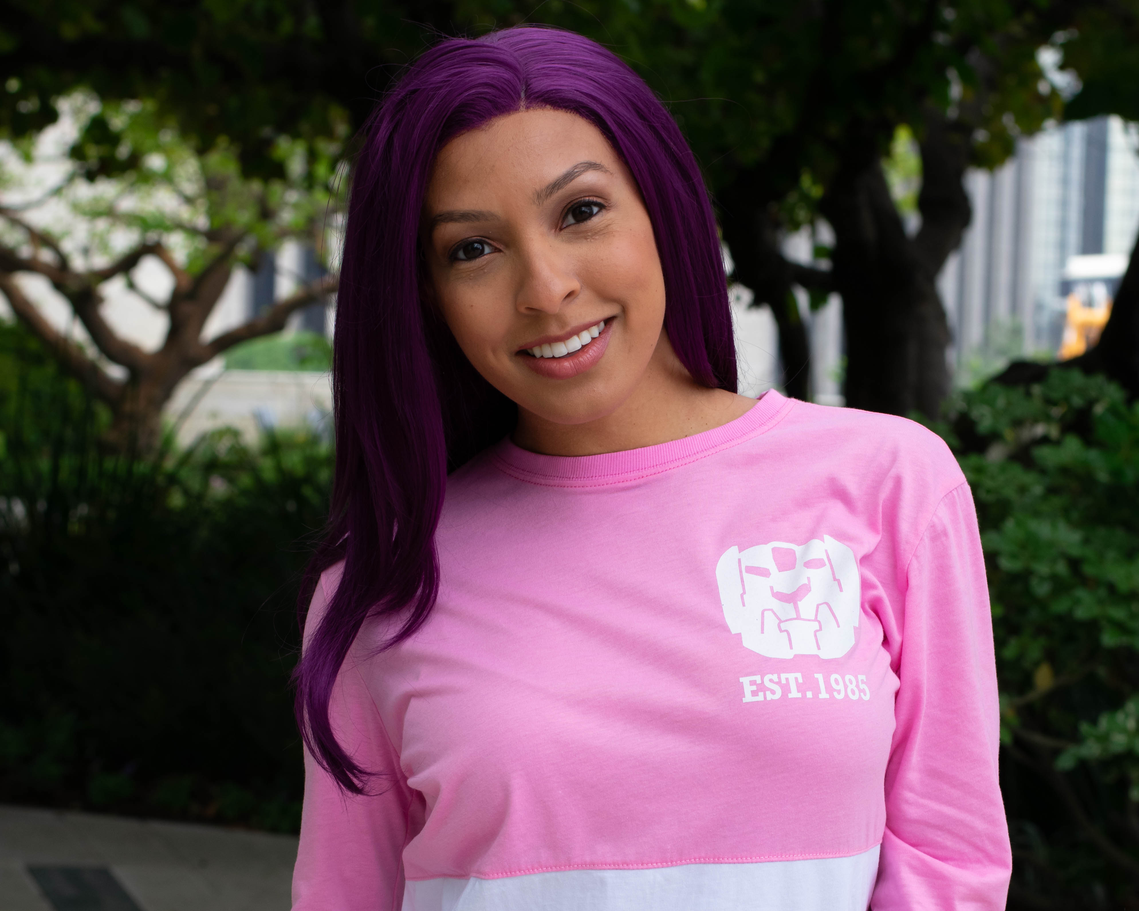 Utahime Cosplay pictured in a pink Voltron shirt. She is standing in a courtyard with trees and bushes behind her