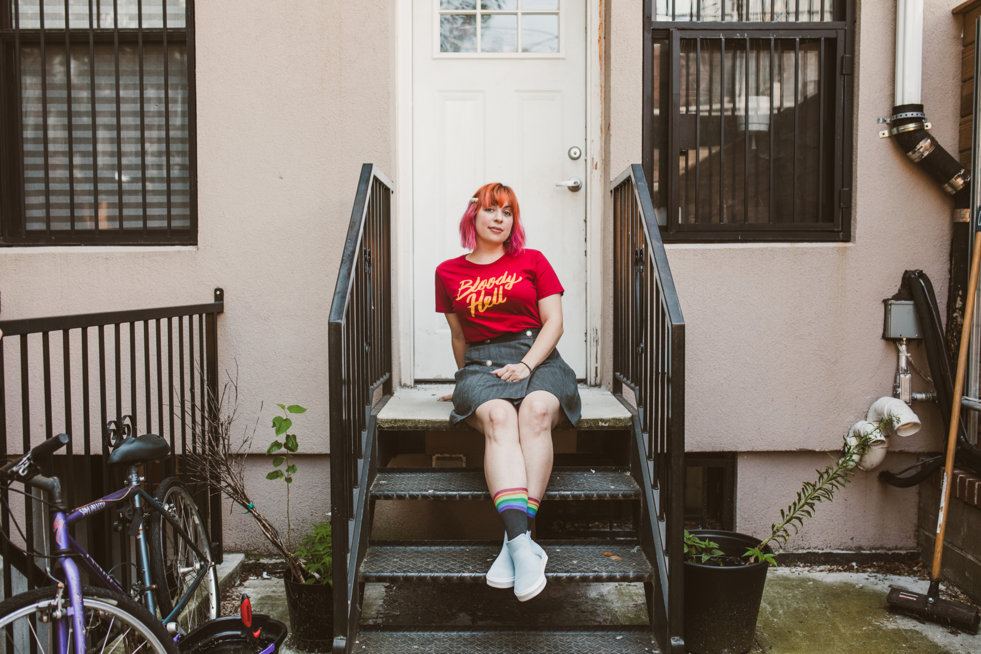 Jordan is sitting on a front porch steps/ Wearing a shirt from her clothing brand. The shirt reads "bloody hell" 