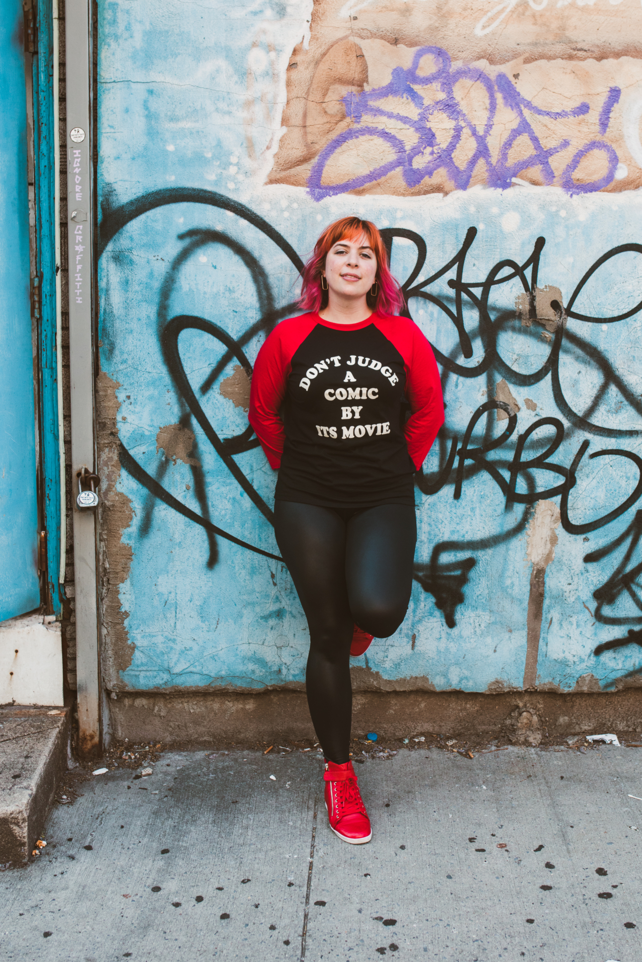 Jordan stands against a wall covered in graffiti. She is wearing a shirt that says "Don't judge a comic by its movie"