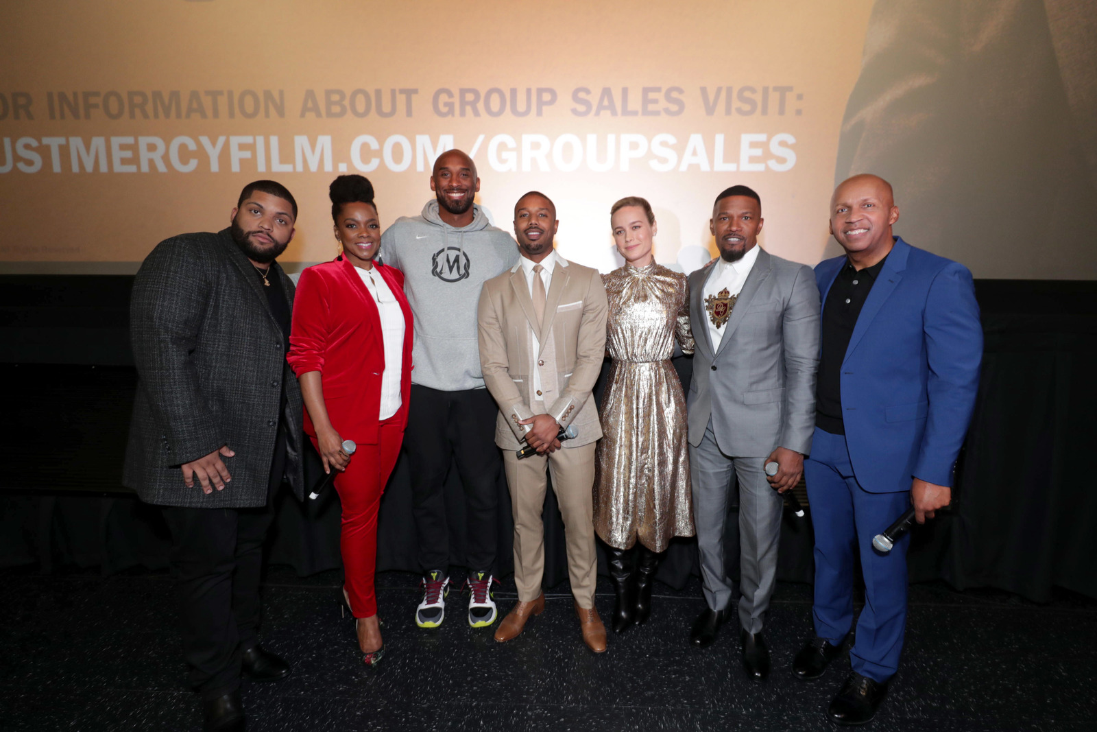 Photo of Michael B. Jordan, Jamie Foxx, Brie Larson, O’Shea Jackson Jr., and Karan Kendrick and author and executive producer Bryan Stevenson attend a special community screening hosted by Kobe Bryant and Terrence J at the Cinemark Baldwin Hills. They are standing in front of the movie screen. 