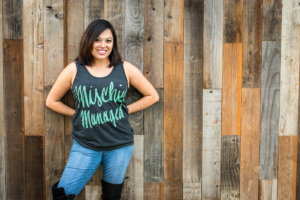 Photo of Monica against a wooden wall wearing a tank top that says "Mischief Managed"