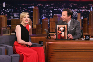 Pictured: (l-r) Actress Jodie Whittaker during an interview with host Jimmy Fallon on January 8, 2020. Jodie is sitting on a couch wearing a black and red dress. Jimmy is wearing a grey suit and is seated behind a desk. Holding a photo of Jodie Whittaker and Mandip gil at the Doctor Who wrap party. both in costumes. Mandip is dressed in Day of the Dead make up and attire. Jodis is in a Kung Fu panda costume.