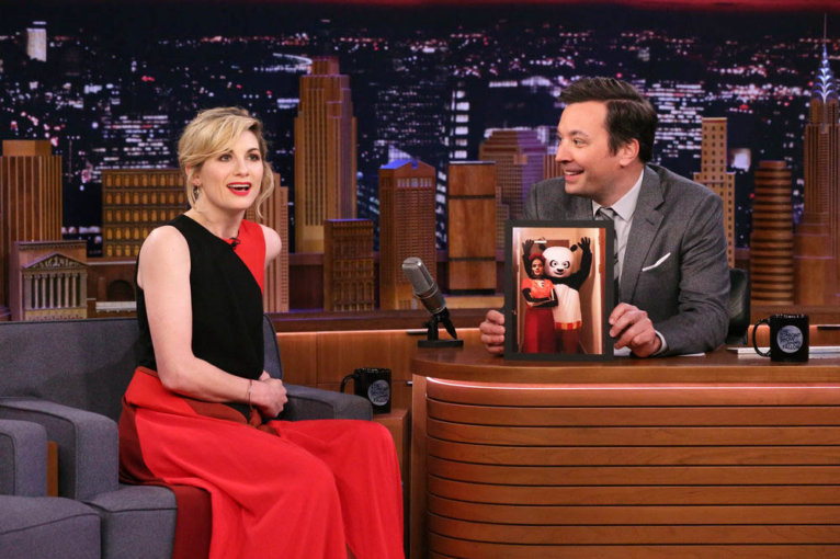 Pictured: (l-r) Actress Jodie Whittaker during an interview with host Jimmy Fallon on January 8, 2020. Jodie is sitting on a couch wearing a black and red dress. Jimmy is wearing a grey suit and is seated behind a desk. Holding a photo of Jodie Whittaker and Mandip gil at the Doctor Who wrap party. both in costumes. Mandip is dressed in Day of the Dead make up and attire. Jodis is in a Kung Fu panda costume.