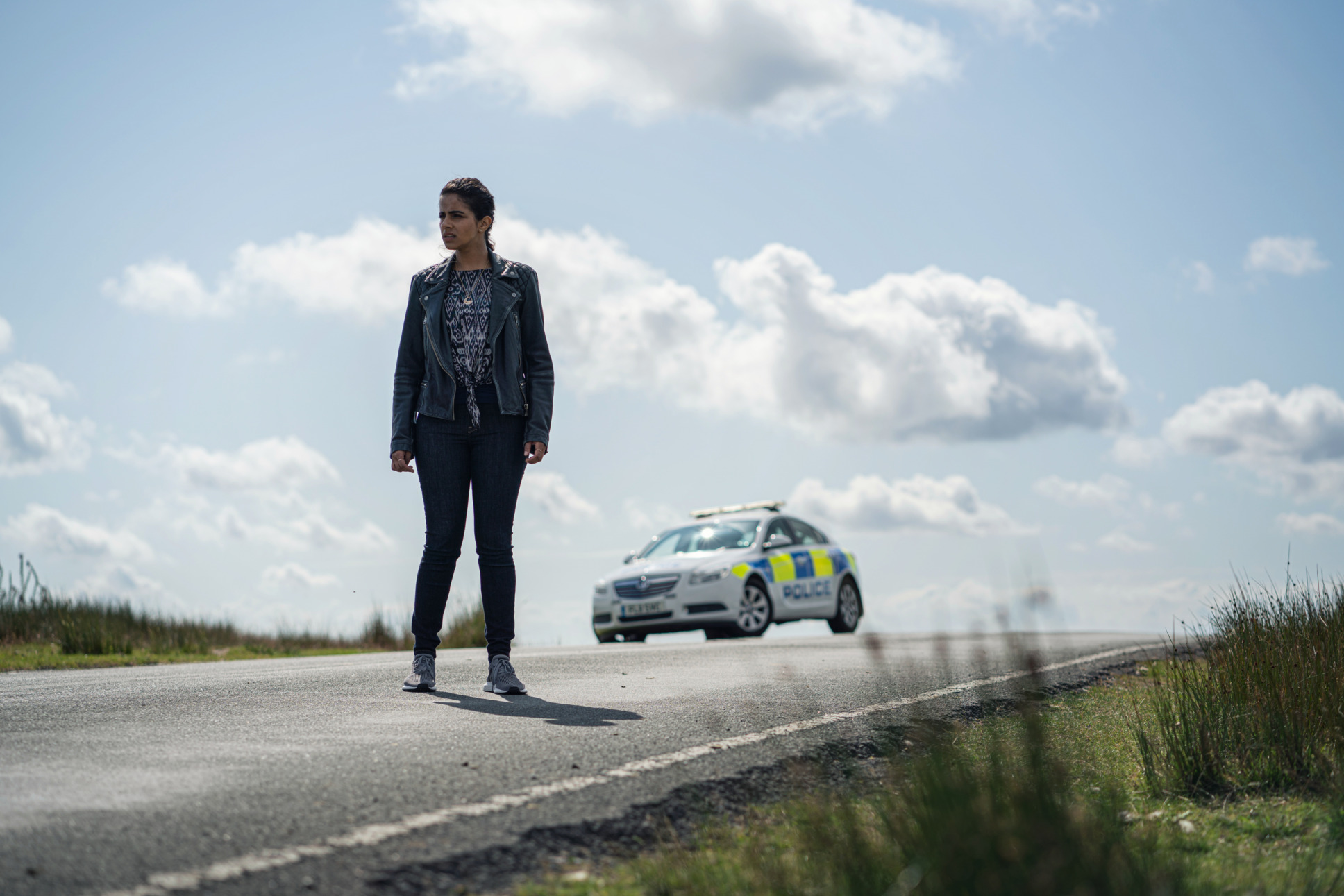 Mandip Gill as Yaz stands on an empty road with only s police car seen in the distance behind her