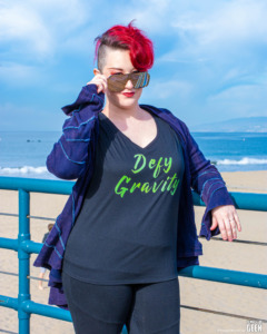 Model is wearning a tshirt that says Defy Gravity. She standing on the pier with the ocean behind her.