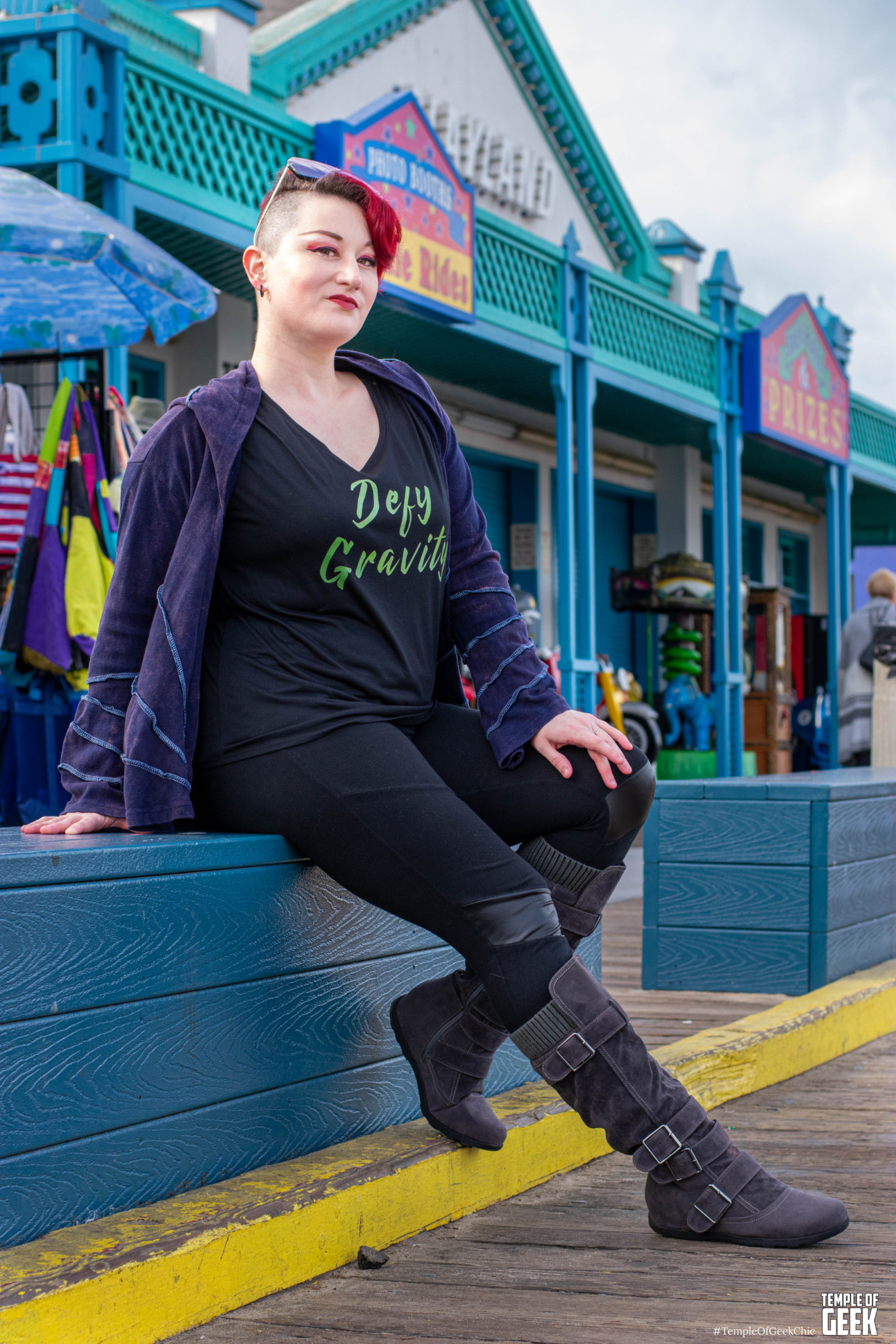 model is sitting on a beach pier in front of a shop wearing a tshirt that says Defy Gravity