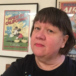 Photo is a headshot of Cynthia Boris. Behind her on the wall is vintage Mikey Mouse art work. 