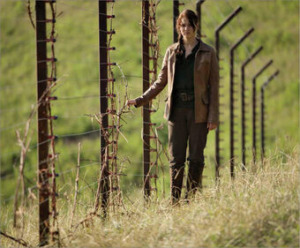 Scene from the Hunger Games. Katniss portrayed by Jennifer Lawrence is walking along the District 12 fence. The fence is covered in barbed wire and the field she is walking on have dried and overgrown grass