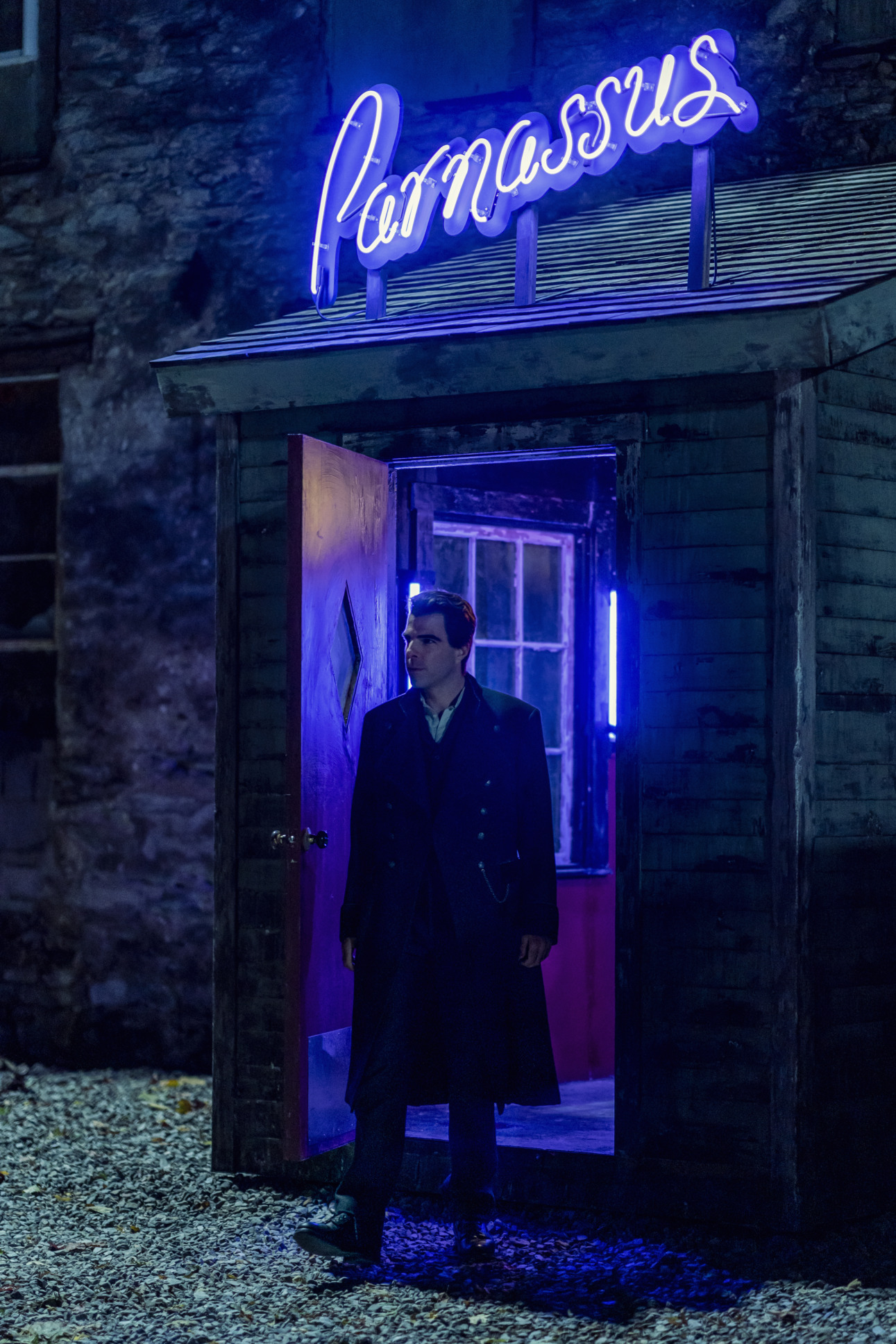 Zachary Quinton as Charlie Manx stands just outside a doorway heading outside. It is nighttime and he is standing under a neon light that reads Parnassus.  Comic Con panel topic