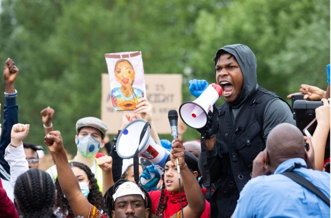 John Boyega speaks in Hyde Park. Boyega is holding a bull horn and is standing on a stool and speaking to protesters gathered around him