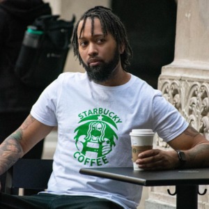 Aaron Powell, Host of the Temple of Geek Podcast sits at an outdoor table on a busy Los Angeles street corner. He holding a Star Bucks coffee cup while wearing a Paradody Winter Soldier Shirt where Bucky is in a Starbucks Logo