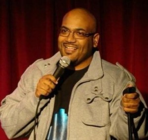 Ed Greer is seen on stage with a microphone infront of him. He is doing stand up comedy