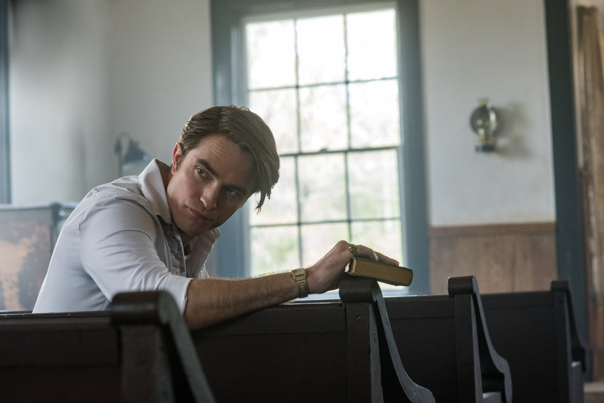 Robert Pattison sits in the pew of a church. His head turned toward the back of the room. He is alone.