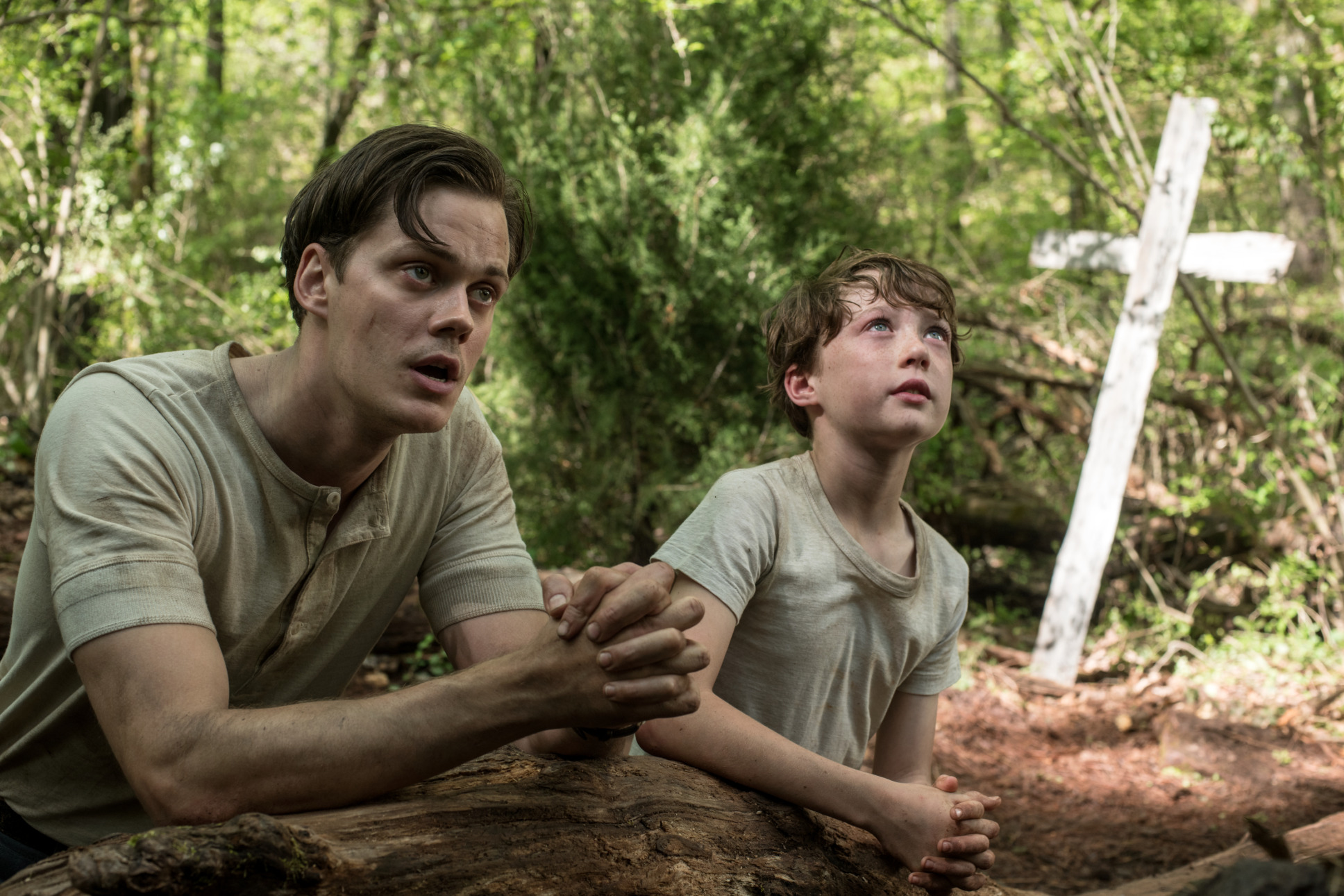 A young man and a young boy sit with hands clasped in prayer in a field. There is a white cross behind them.