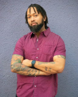 Photo of Podcast Host Aaron Powell standing against a wall with his arms crossed and a smirk on his face. 