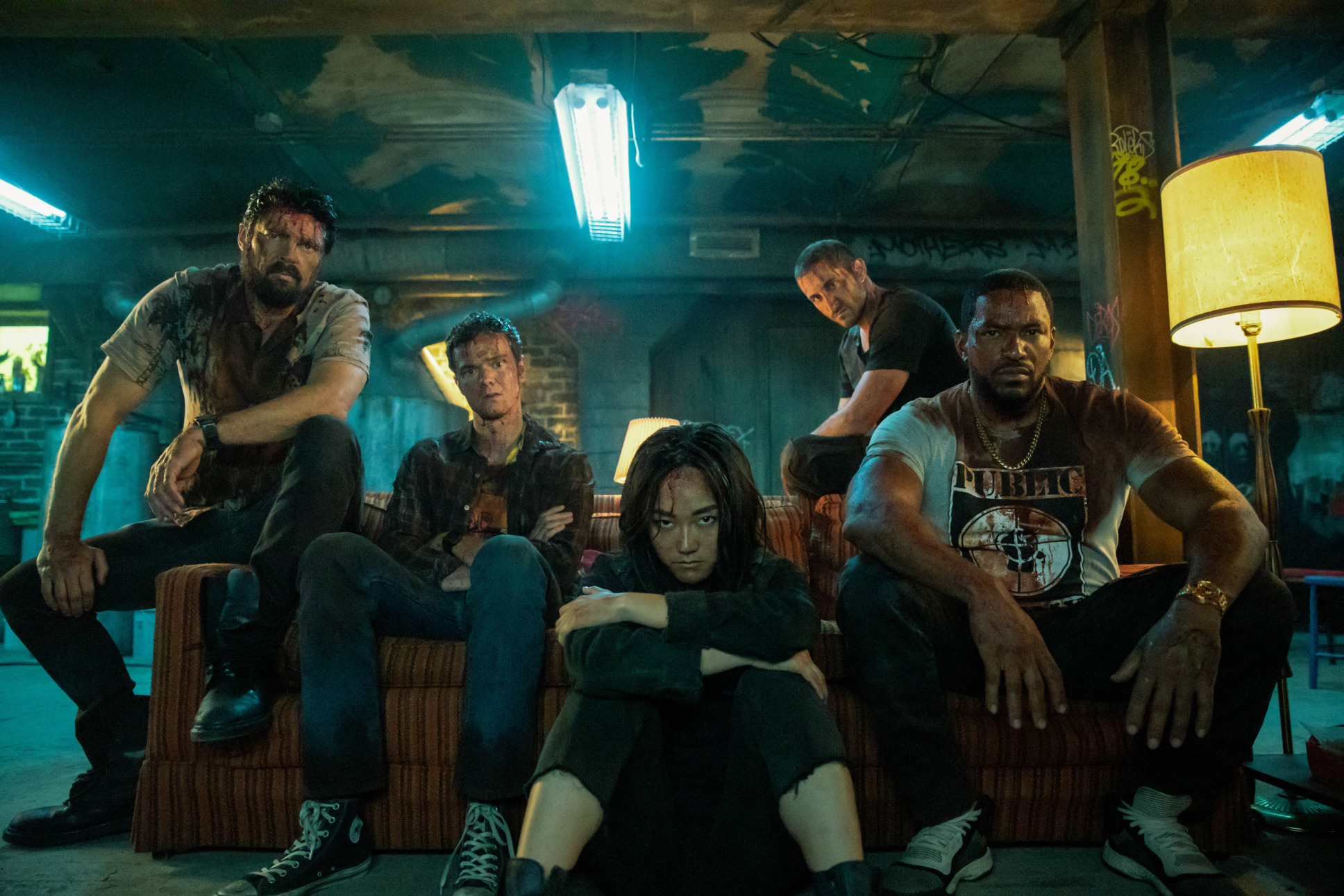 In hiding in a low lit basement, sitting on a couch, Hughie (Jack Quaid), Mother’s Milk (Laz Alonso), Frenchie (Tomer Capon), and Kimiko is also known as The Female (Karen Fukuhara) are staring directly at the camera, covered in dirt and blood. 