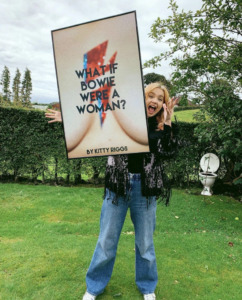 Kitty Riggs holds up a framed photo of her book cover. she is standing in a field