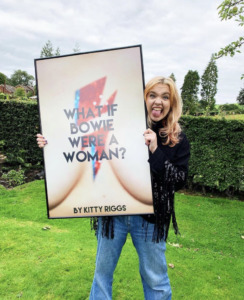 Kitty Riggs sticks her tounge out as she poses for a photo. She is holding up a photo of her book cover