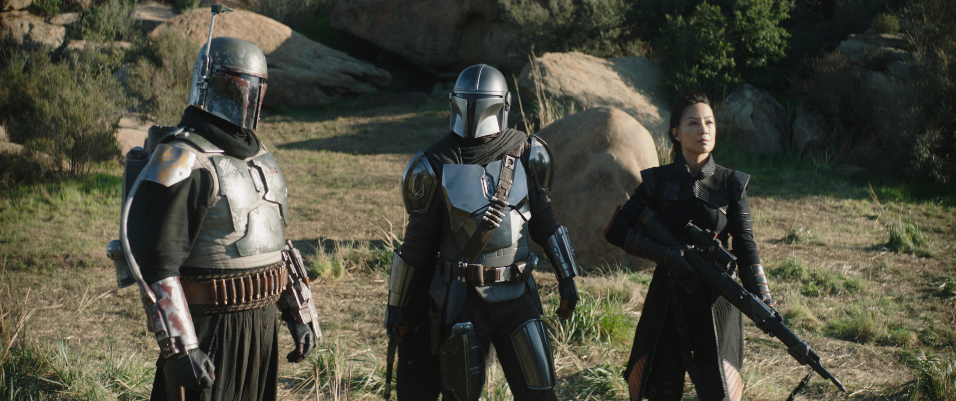 Boba Fett, the Mandalorian, and Fennec Shand are standing in the desert in front of some rocks