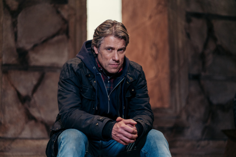 Picture Shows: Dan (JOHN BISHOP) sitting on the steps in front of a rocky stone monument. and opening appears behind him with light shinning through. possibly a door way. 