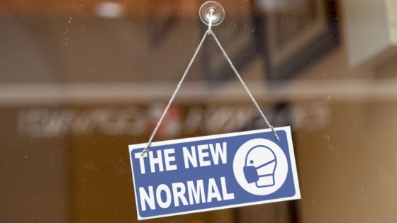 A since hangs on a shop window. The sign says "the new normal" and has agraphic of a person wearing a facemask