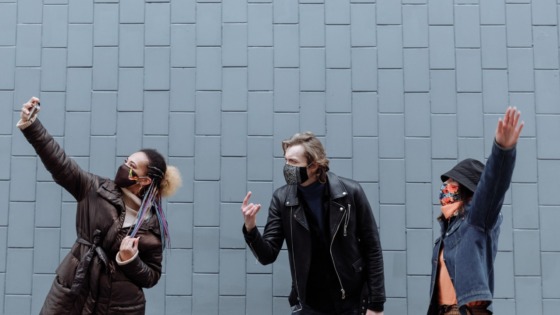 Man in Black Leather Jacket Standing Beside Woman in Black Leatherand woman in a demin jacket, all wearing facemasks. They are posing for a selfie while separated by several inches