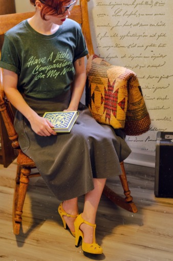 Ashley Rose sits on a chair in ger bedroom. She is holding a book and wearing a t-shirt by Jordandene that she has styled with a pair of oxford heels and a mid length skirt. 