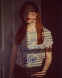 Ashley Rose faces a window and a shadow from the window blinds is cast across her face. wearing a t-shirt by Jordandene 