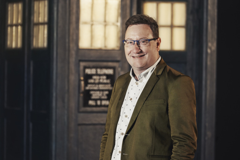 Chris Chibnall smiles at the camera as he stands in front of the TARDIS