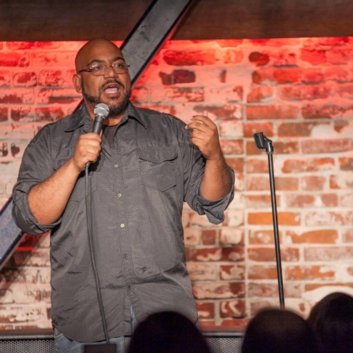 Ed Greer is on stage at a comedy club holding a microphone and looking out and speaking to a dark crowd infront of him. 