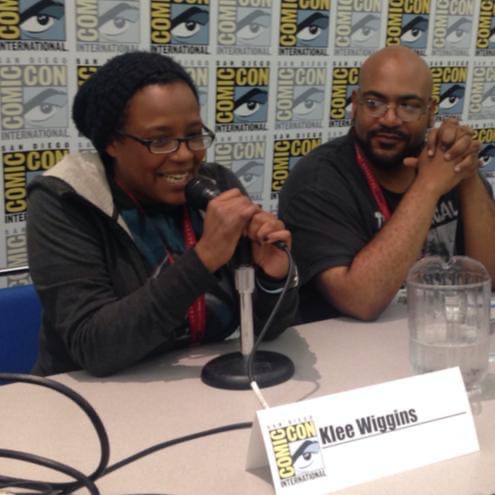 Klee Wiggins and Ed Greer at San Diego Comic Con, sitting at a panel table, klee is talking into a microphone addressing an unseen crowd. Ed is clasping his hands as he looks over and listens to Klee