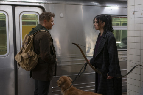 Kate Bishop, holding her bow and a leash to Lucky the Pizza Dog,, stands face to face with Clint Barton next to an underground subway train.