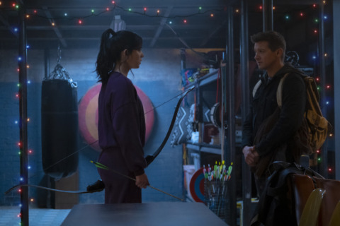 Kate Bishop, holding her bow, stands face to face with Clint Barton in a room of random stuff.