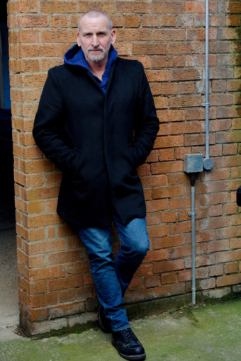 a picture of actor Christopher Eccleston dressed in a black coat and blue jeans, leaning against a brick wall