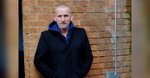 a picture of actor Christopher Eccleston dressed in a black coat and blue jeans, leaning against a brick wall