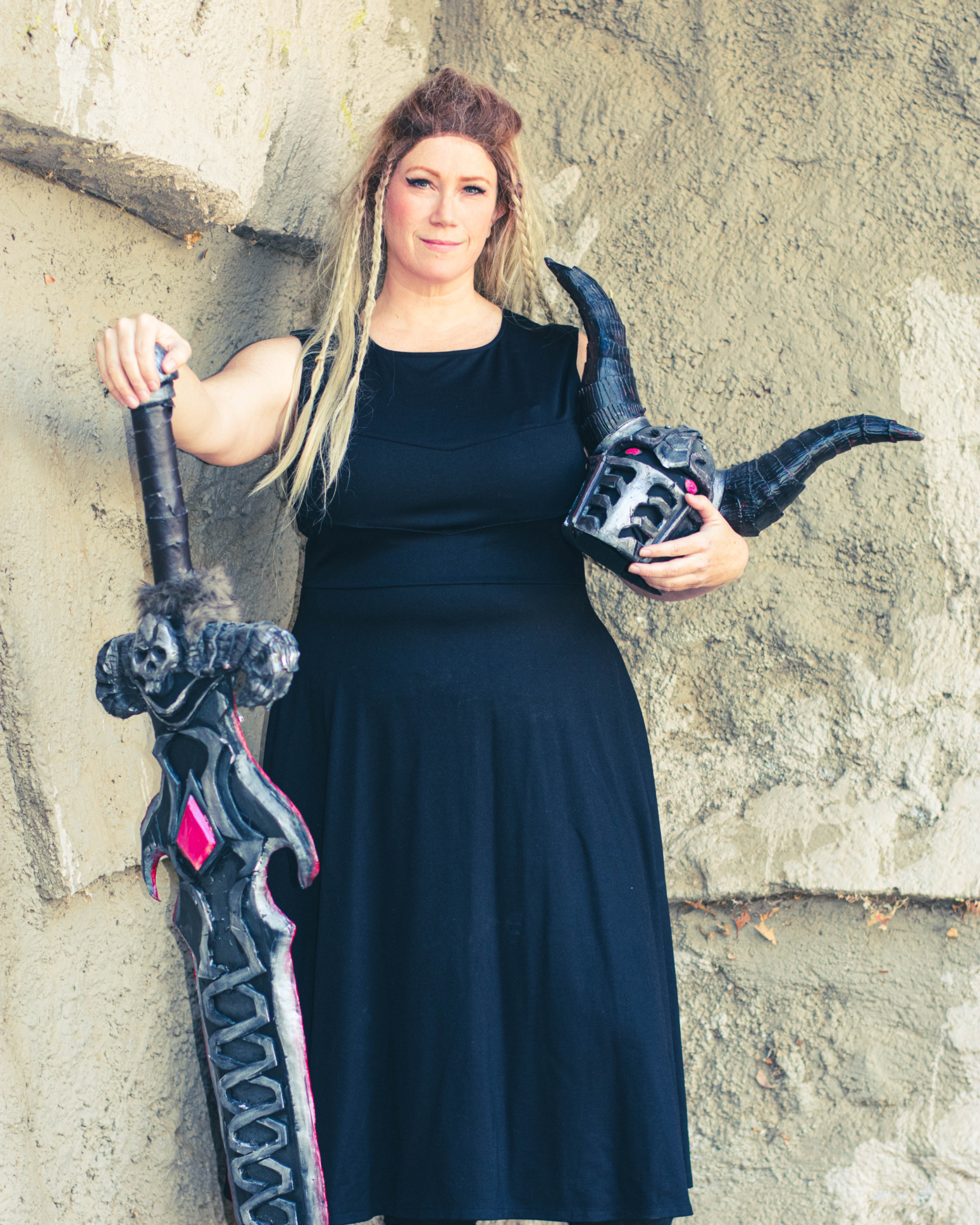 A model holding a helmet with horns and a sword models a black Longsword dress from D&D fashion brand Guild Party.