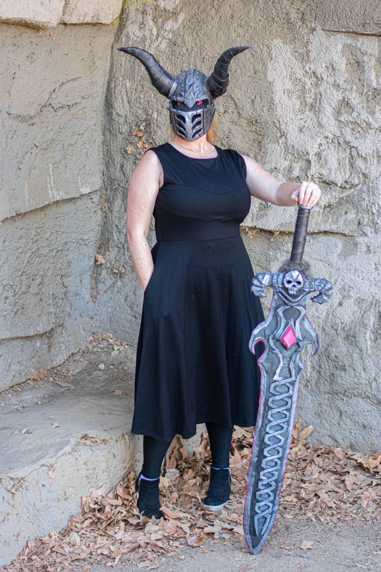 A model wearing a helmet with horns and holding a sword models a black Longsword dress.