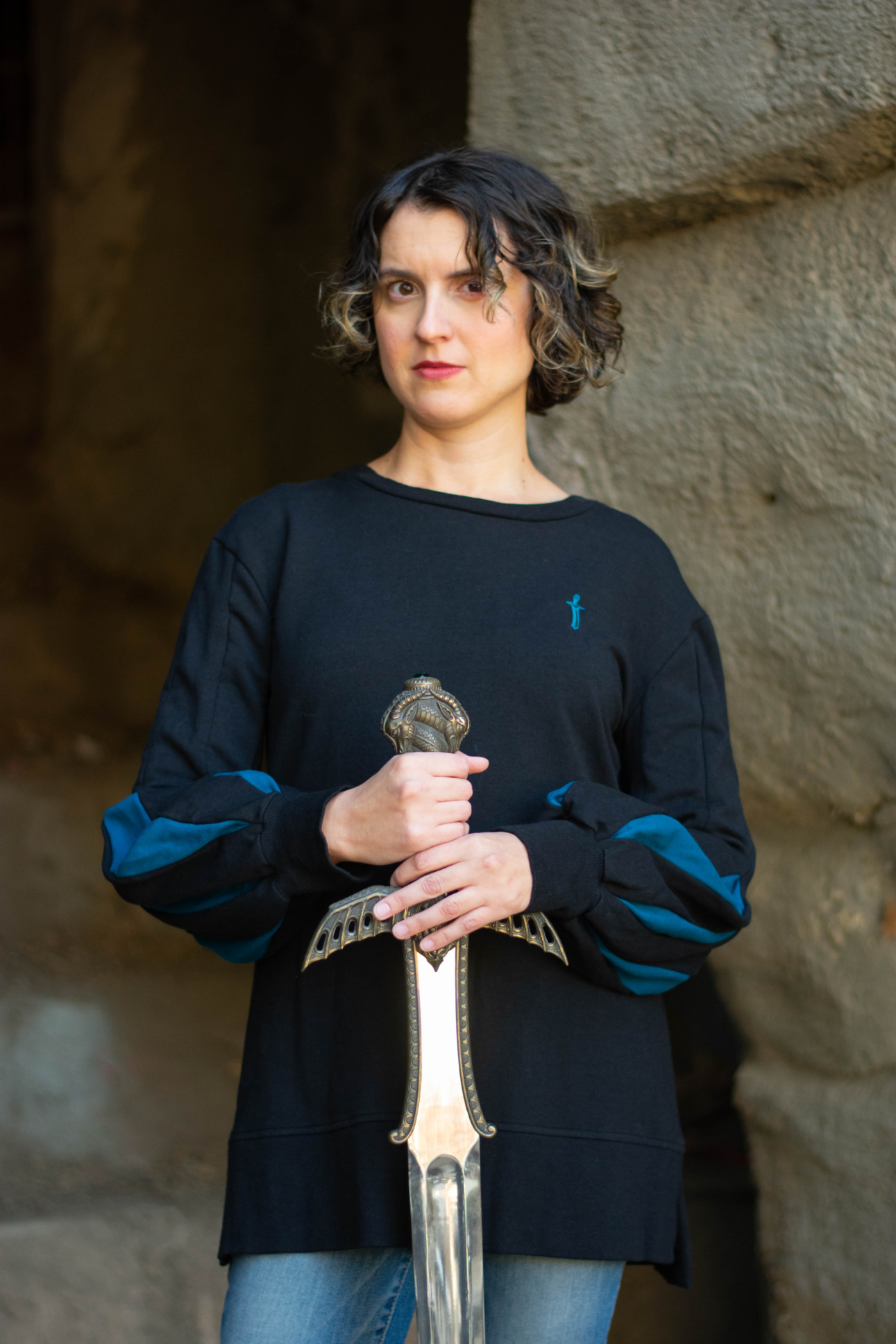 A model poses with a sword and wears a black pullover featuring bishop sleeves and an embroidered sword emblem.