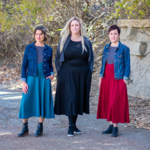 Three models wear denim jackets over teal, black, and ruby dresses from D&D fashion brand Guild Party.