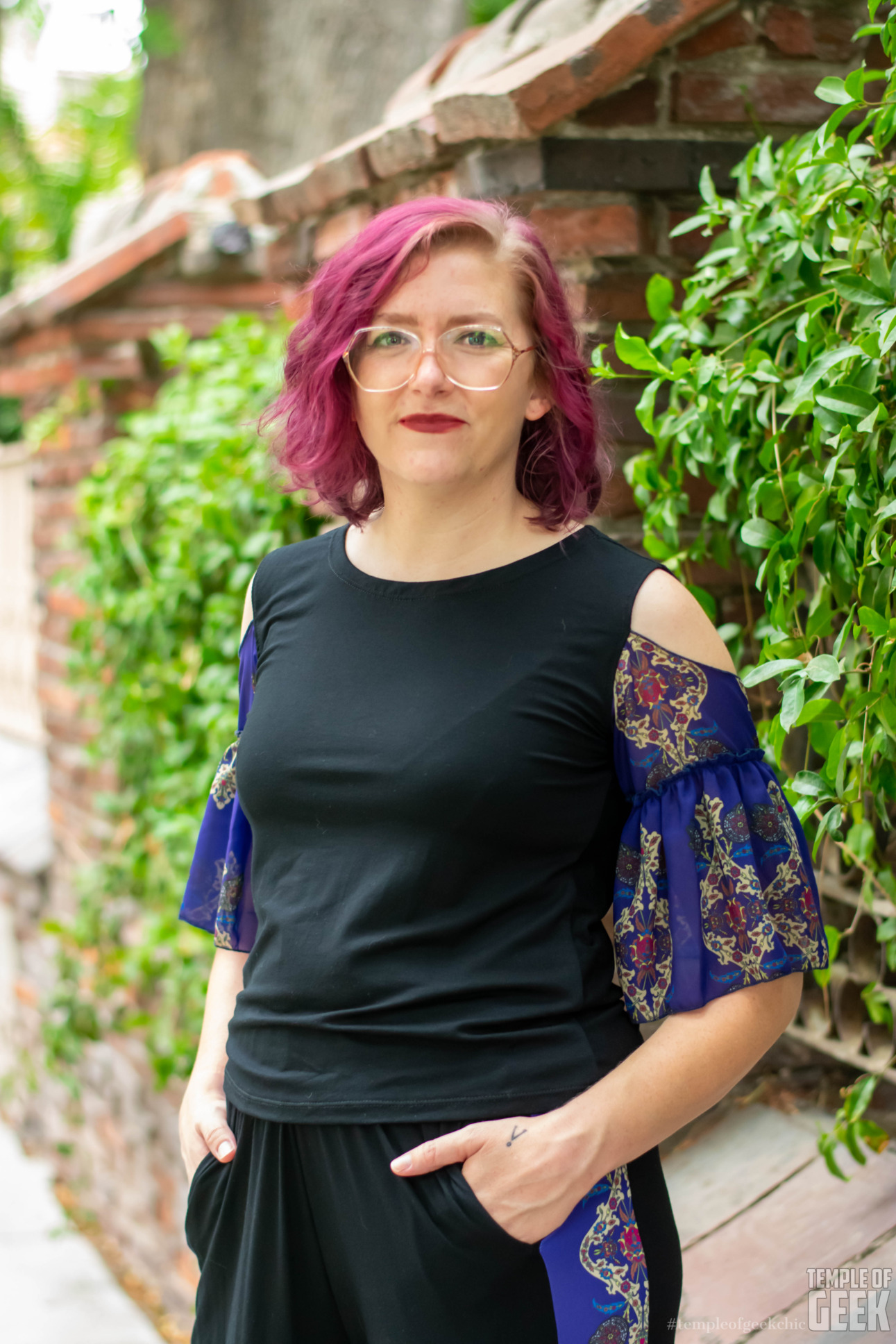 A model stands in front of foliage wearing a deep blue ruffle sleeve top by Heroicouture.