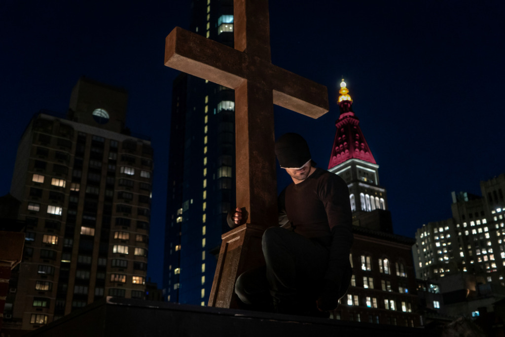 Charlie Cox as Daredevil sitting perched on a roof ledge next to a standing cross