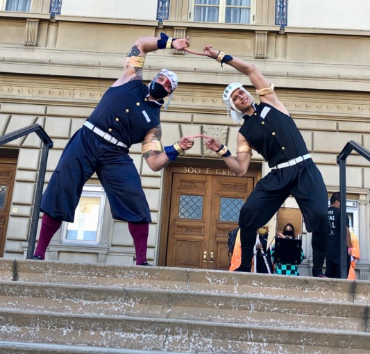 two Tengen cosplayers touch pinkie fingers while forming a circle with their arms as they pose for a photo