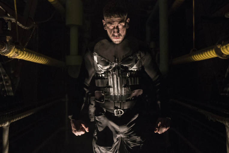 The Punisher played by Jon Bernthal standing in a hallway with The Punisher embem on his chest