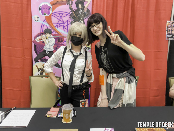 Maddie Morrow and Kayleigh McKee at Anime Riverside 2022. They are standing at the exhibitor booth for Kayleigh. Kayleigh is holding up a peace sign and smiling. Maddies is doing the same, her smile is covered by a facemask.