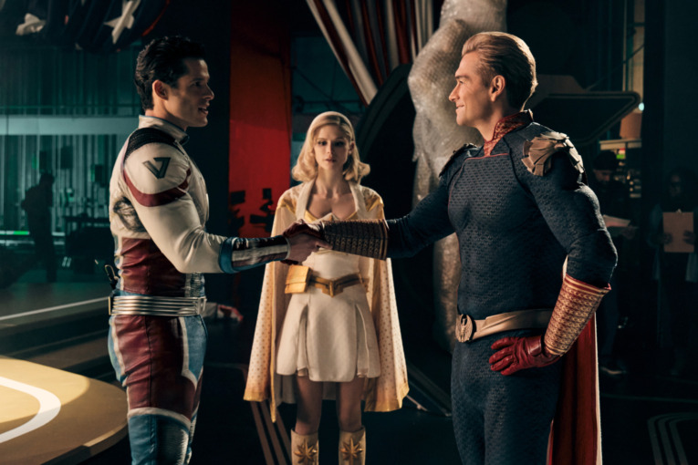 (L-R) Miles Gaston Villanueva as Super Sonic, Erin Moriarty as Starlight and Antony Starr as Homelander. SuperSonic and Homelander shake hand and face one another as if to signify that they have made a deal. Starlight stands behind them with her arms crosses as if she is upset by the arrangement. 