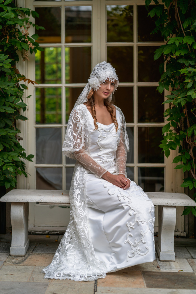 A model sits on a bench in front of a window surrounded by greenery while wearing a lacy white veil and replica of a wedding dress and lacy veil from Star Wars: Attack of the Clones. 