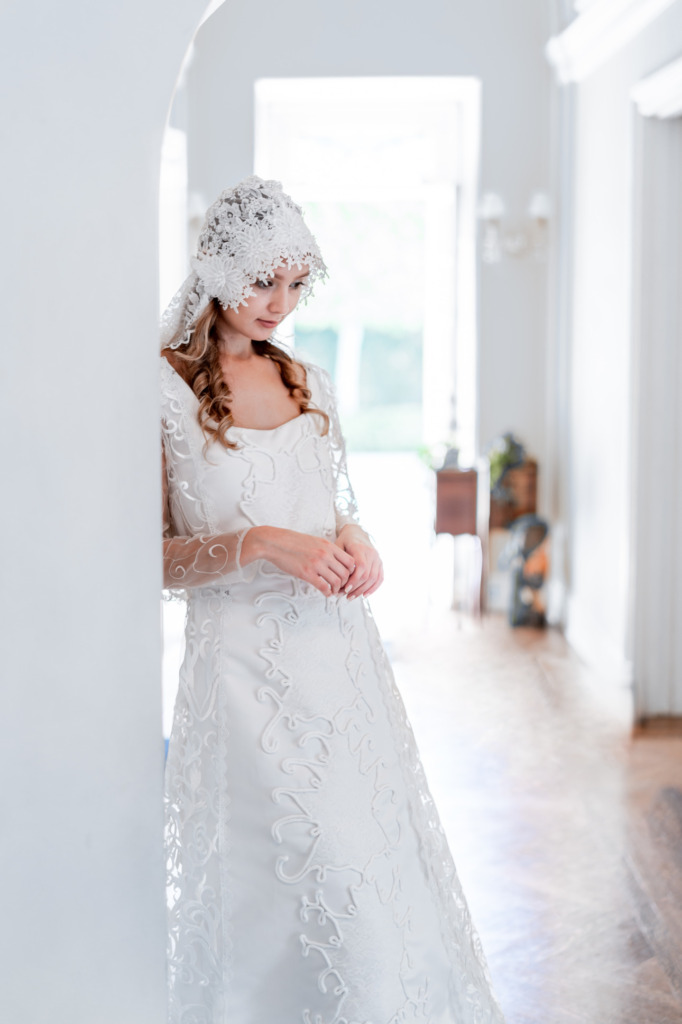 A model leans against a wall in a white room filled with light while wearing a replica of Padme Amidala's wedding dress and lacy veil from Star Wars: Attack of the Clones. 